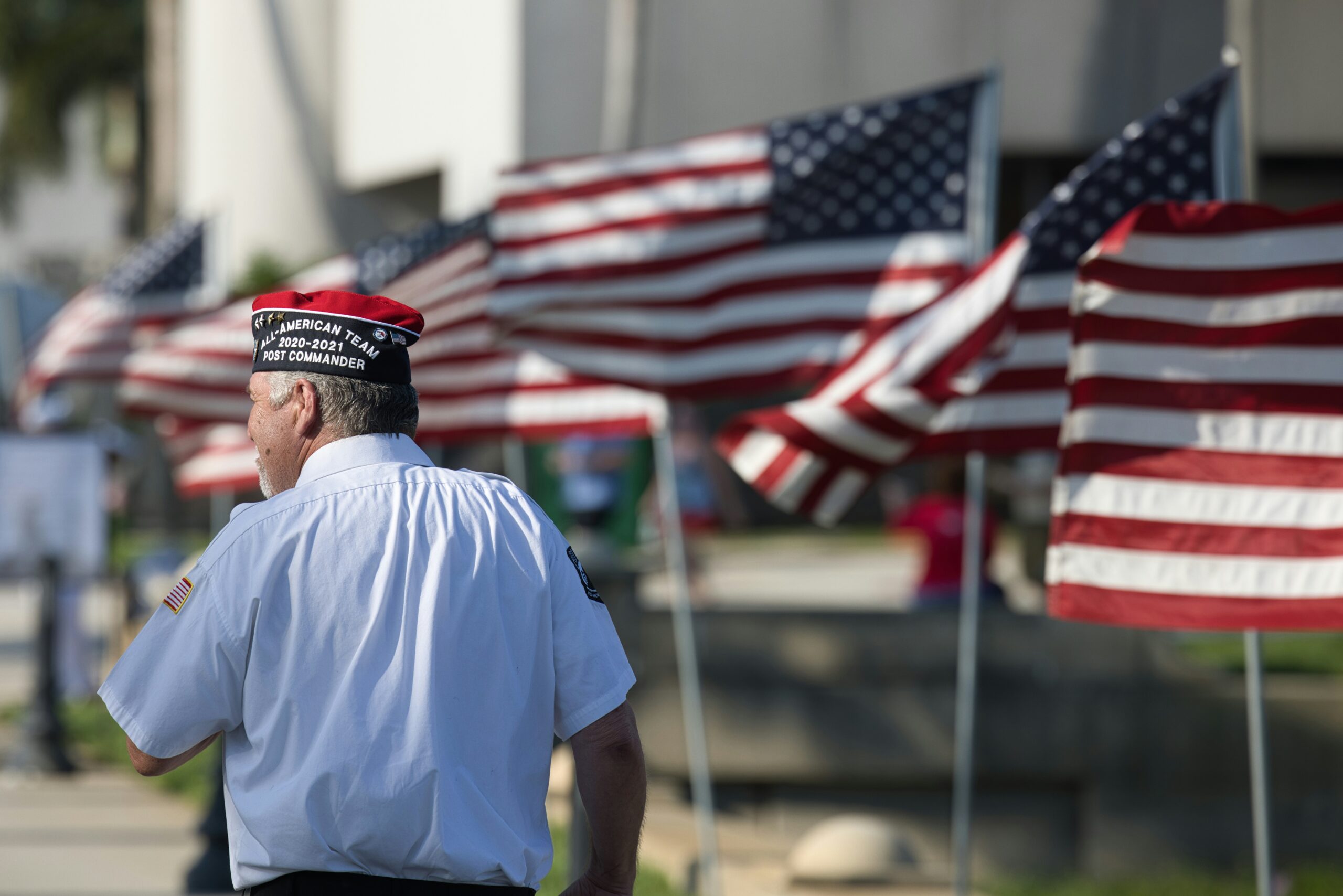 An older veteran carrying a large American flag over his shoulder, walking past a display of multiple flags outdoors.